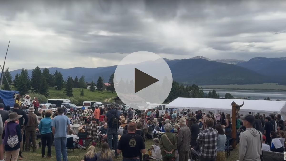 A still image with a play button on top shows a large group of people gathered outside with green trees and tall mountains in the distance