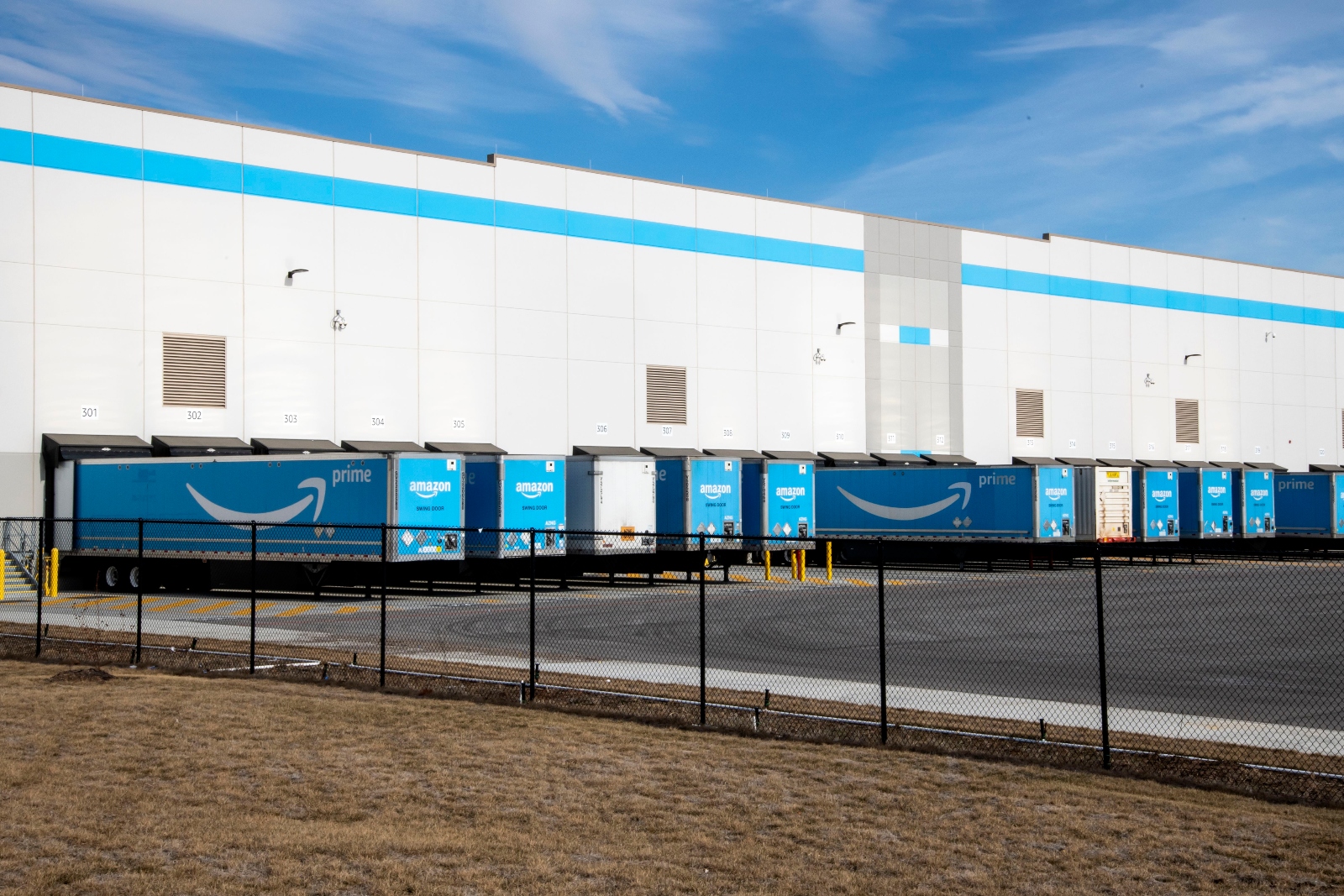 The outside of a white warehouse building with trucks bearing Amazon Prime's branding and logo lined to load from it