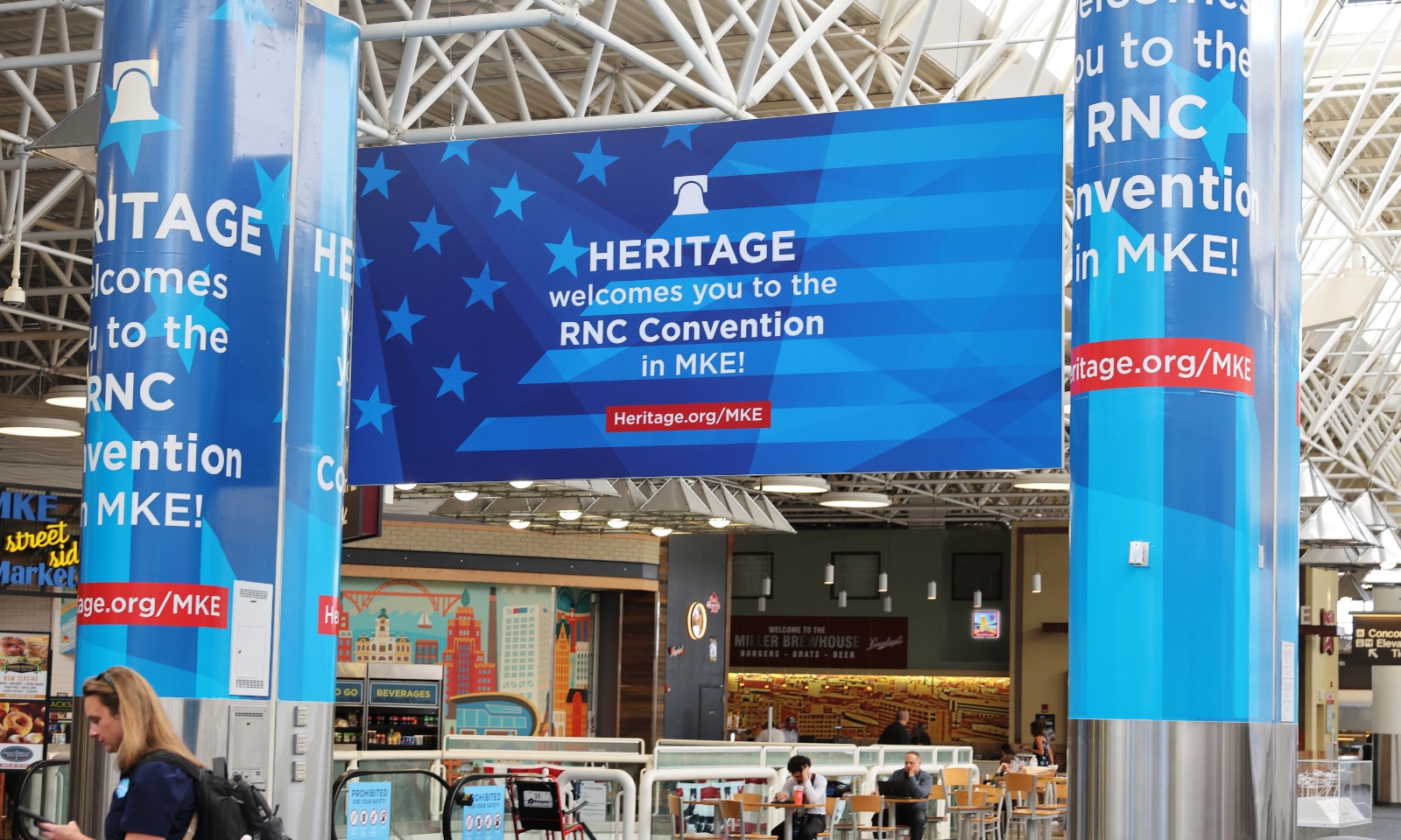 A large, blue sign with the words "Heritage welcomes you to the RNC Convention in MKE" on it hangs between two pillars at the Milwaukee airport.