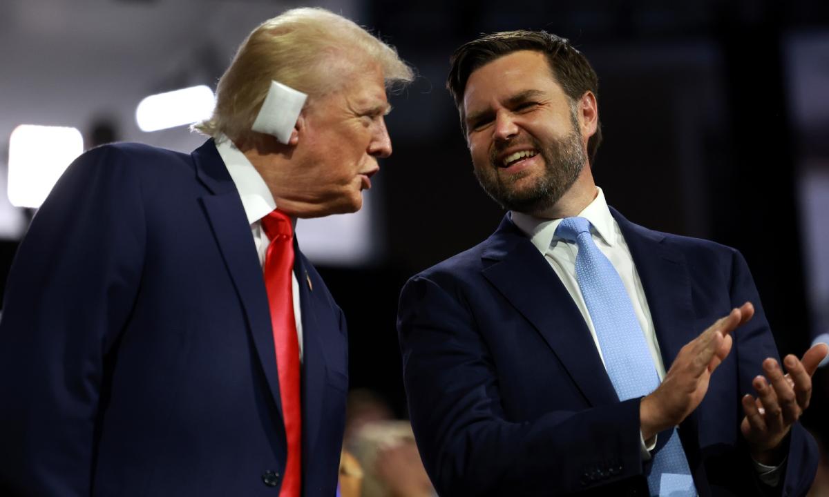 Donald Trump and J.D. Vance, wearing suits, stand on stage. Trump, wearing a white bandage on his ear, is saying something to Vance.