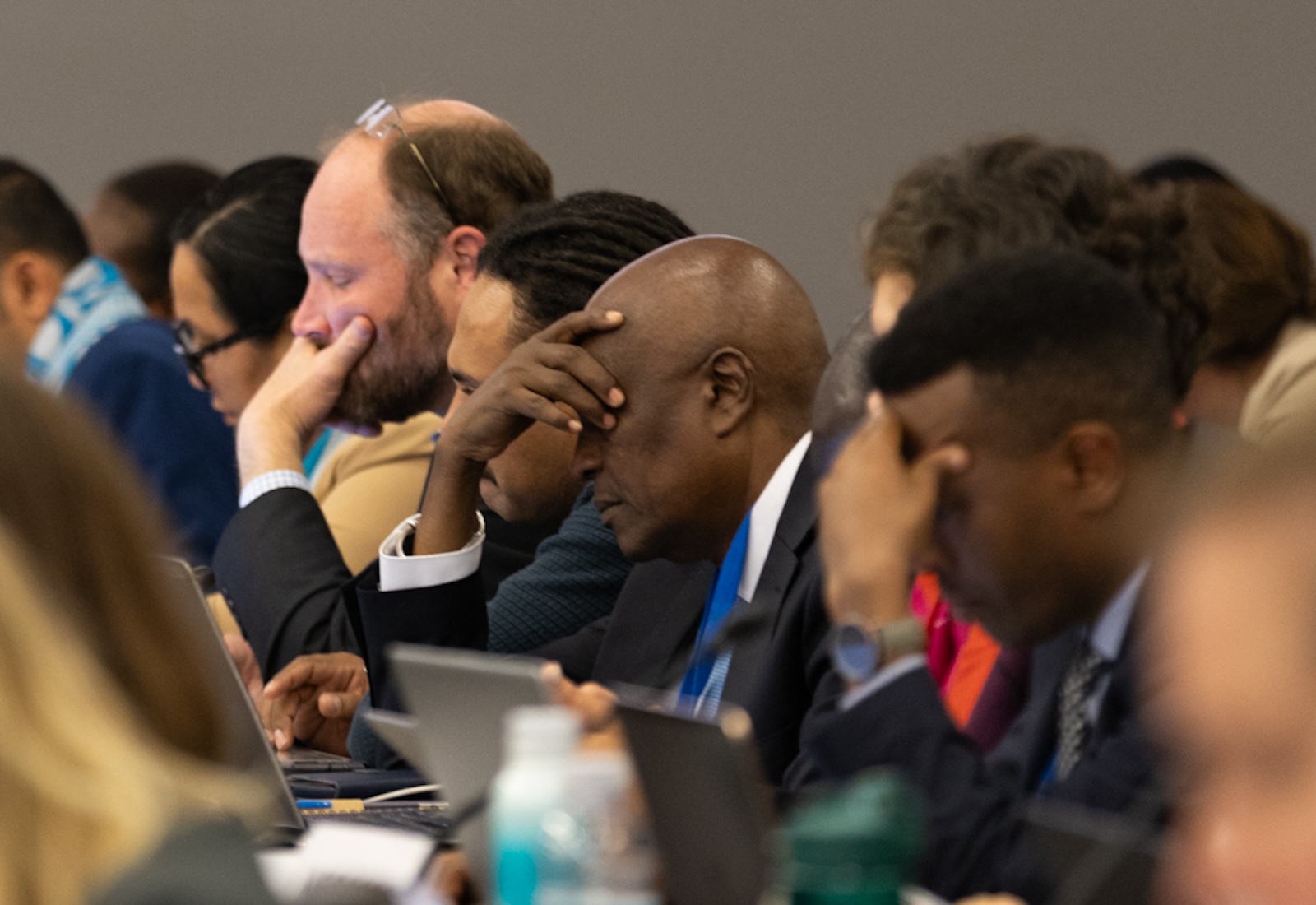 People seated at a table looking at laptops. A man in the center has his hand covering his eyes and appears to be deep in thought.