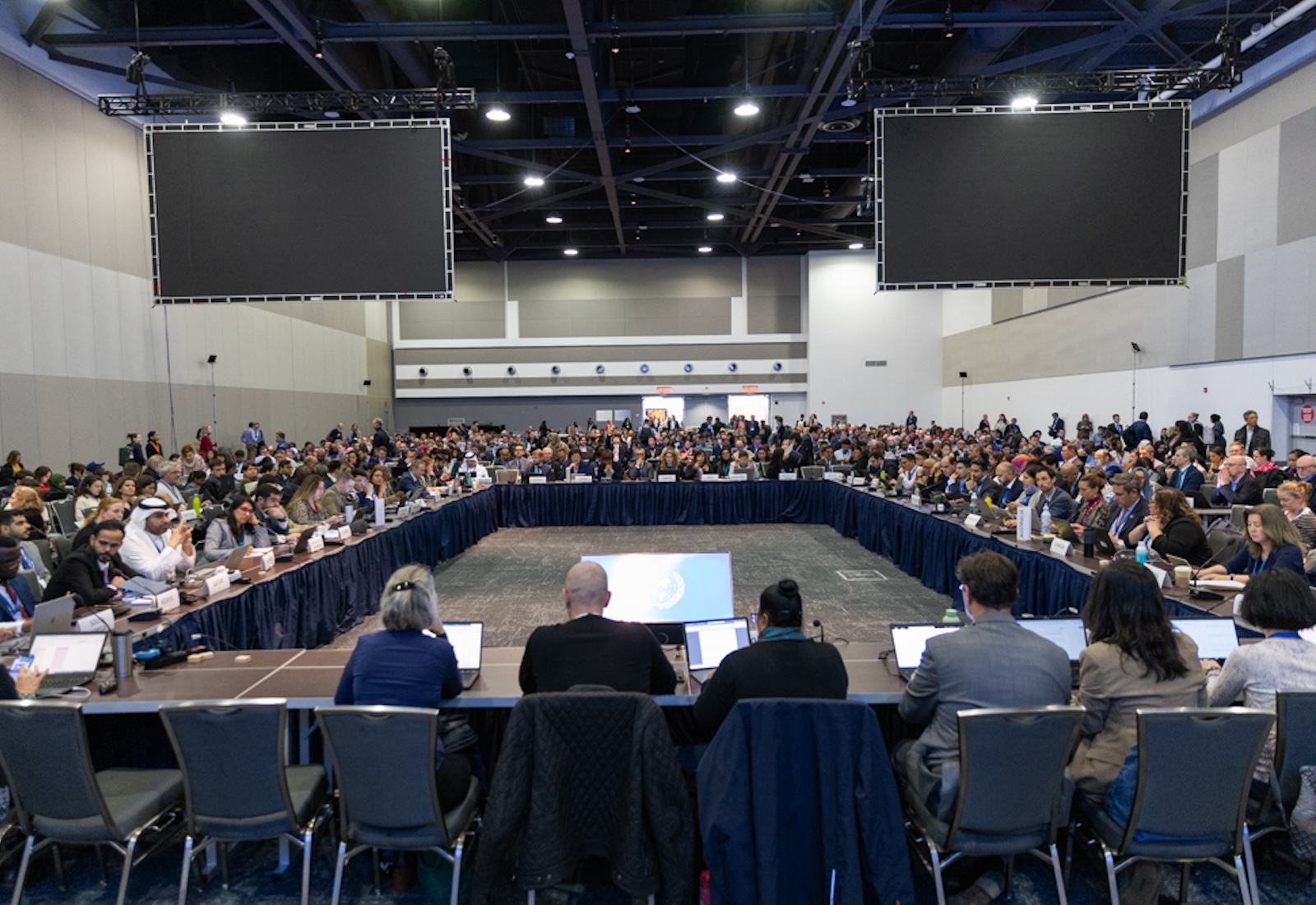 Delegates sit in at tables arranged in a rectangle, with blank screens lifted in the air above them.