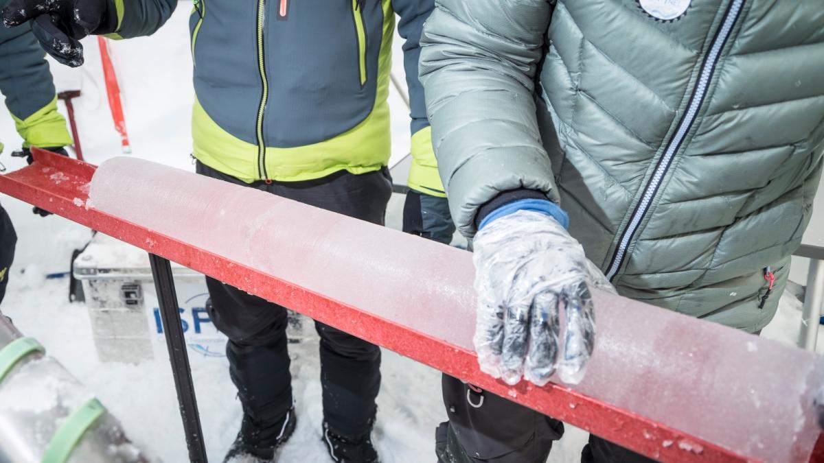 People in parkas and thick gloves handle a cylinder of ice sitting on a thin red platform