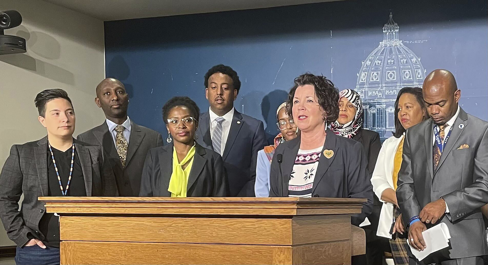 A group of people stand in front of a podium. In the center, speaking, is a woman in a suit.