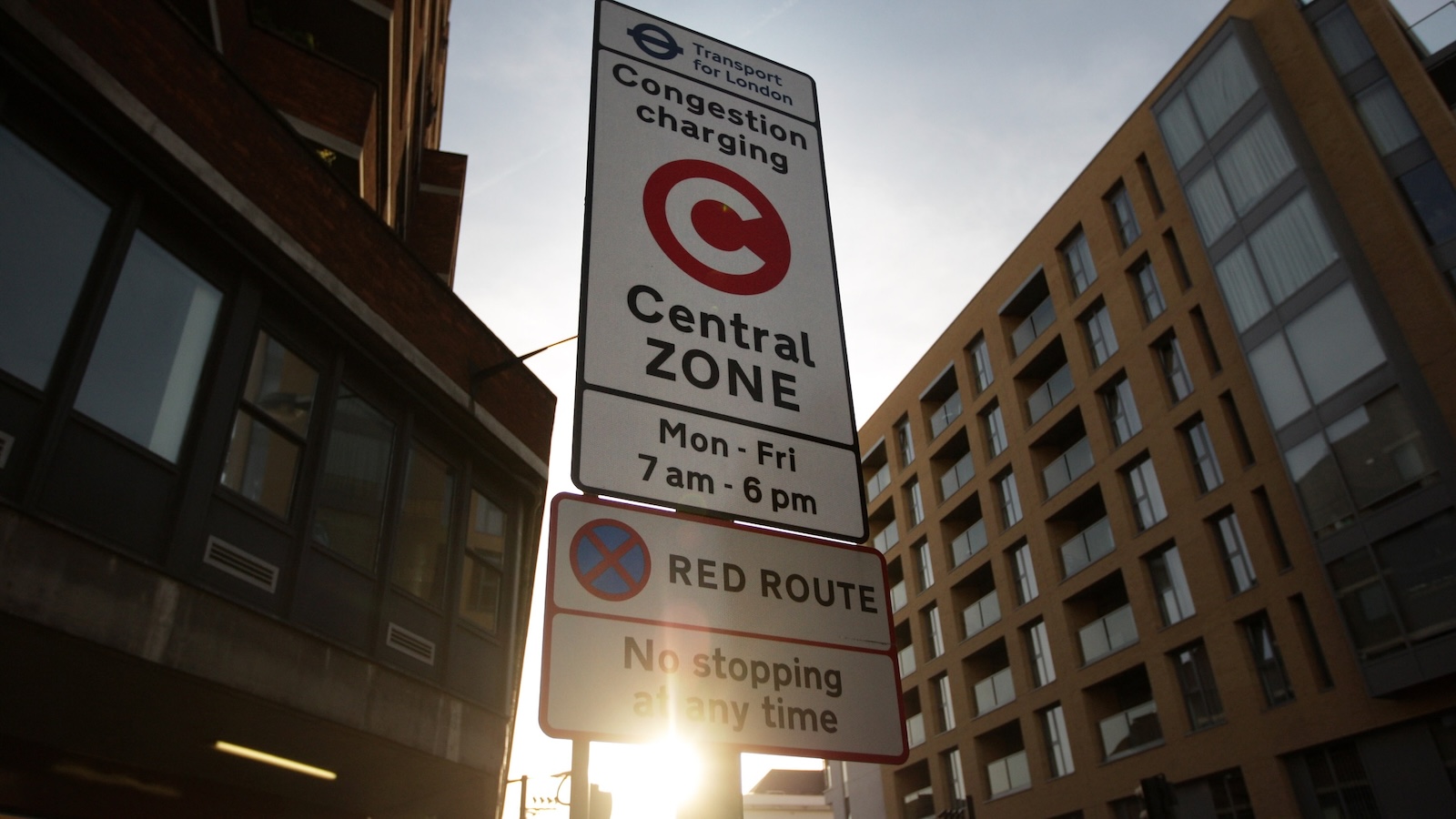 A sign, framed by the buildings on either side of it, informs motorists that they are entering London's congestion charge zone. The sign reads "Congestion charging central zone. Monday through Friday, 7 am through 6 pm."