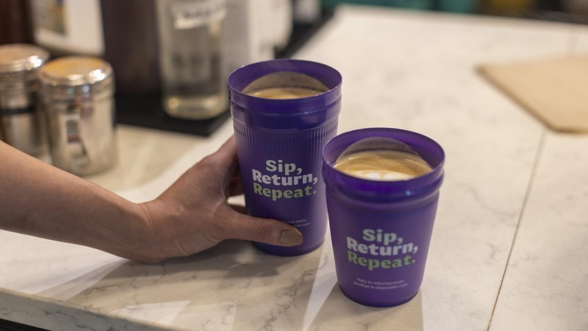 Two purple cups filled with coffee. A hand holds one. Both read, "Sip, Return, Repeat" and are set on a wooden table.