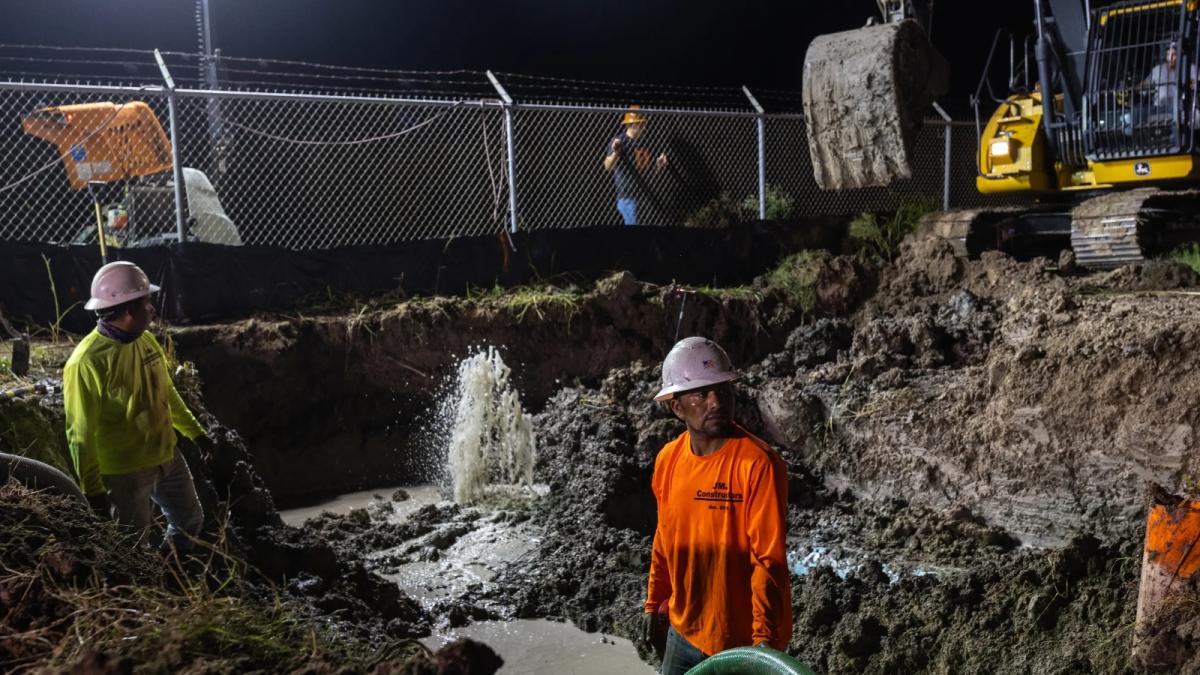 A group of construction workers stand in a trench at night.