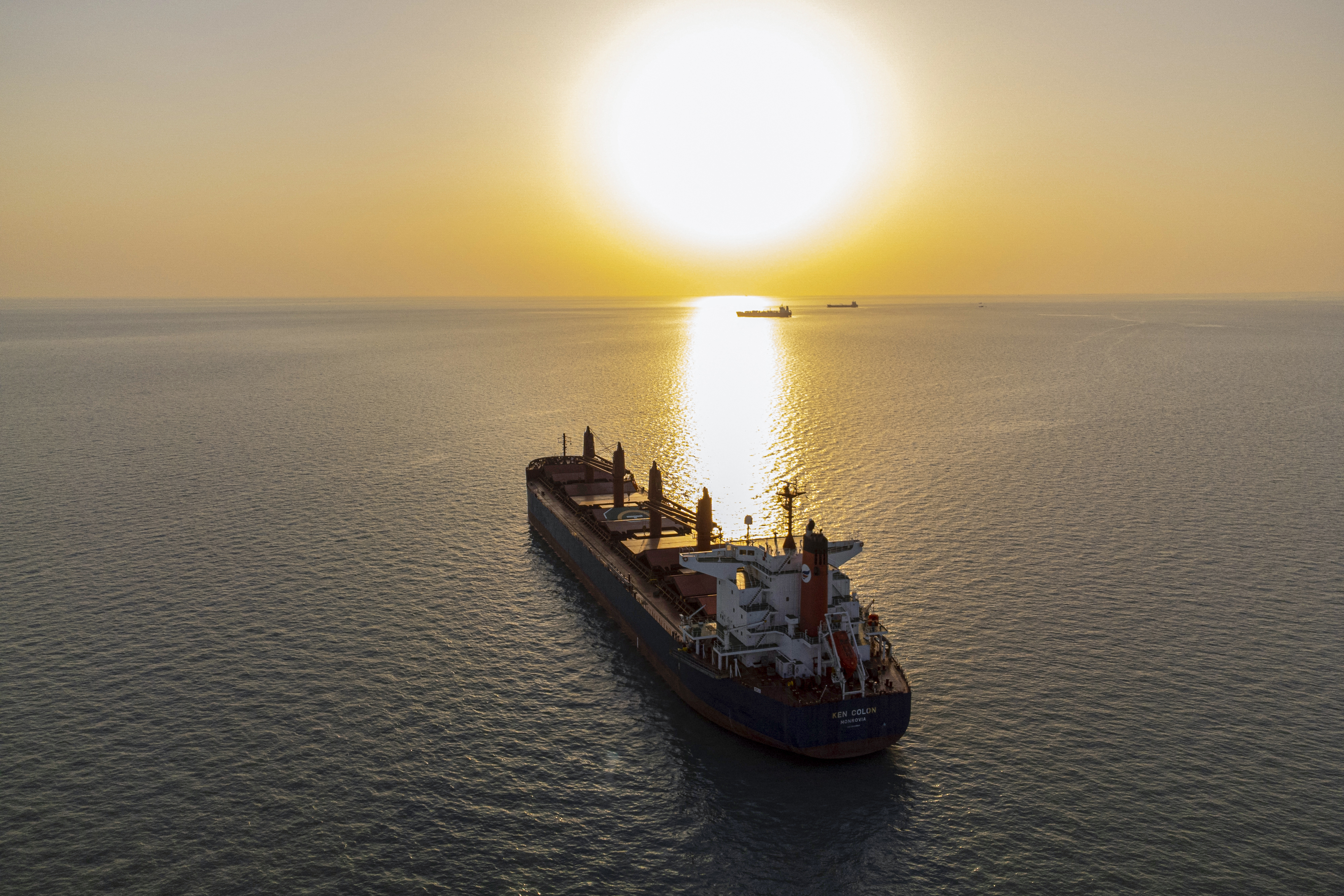 A cargo ship rests on the open ocean as the sun sets behind it, glinting off the sea surface