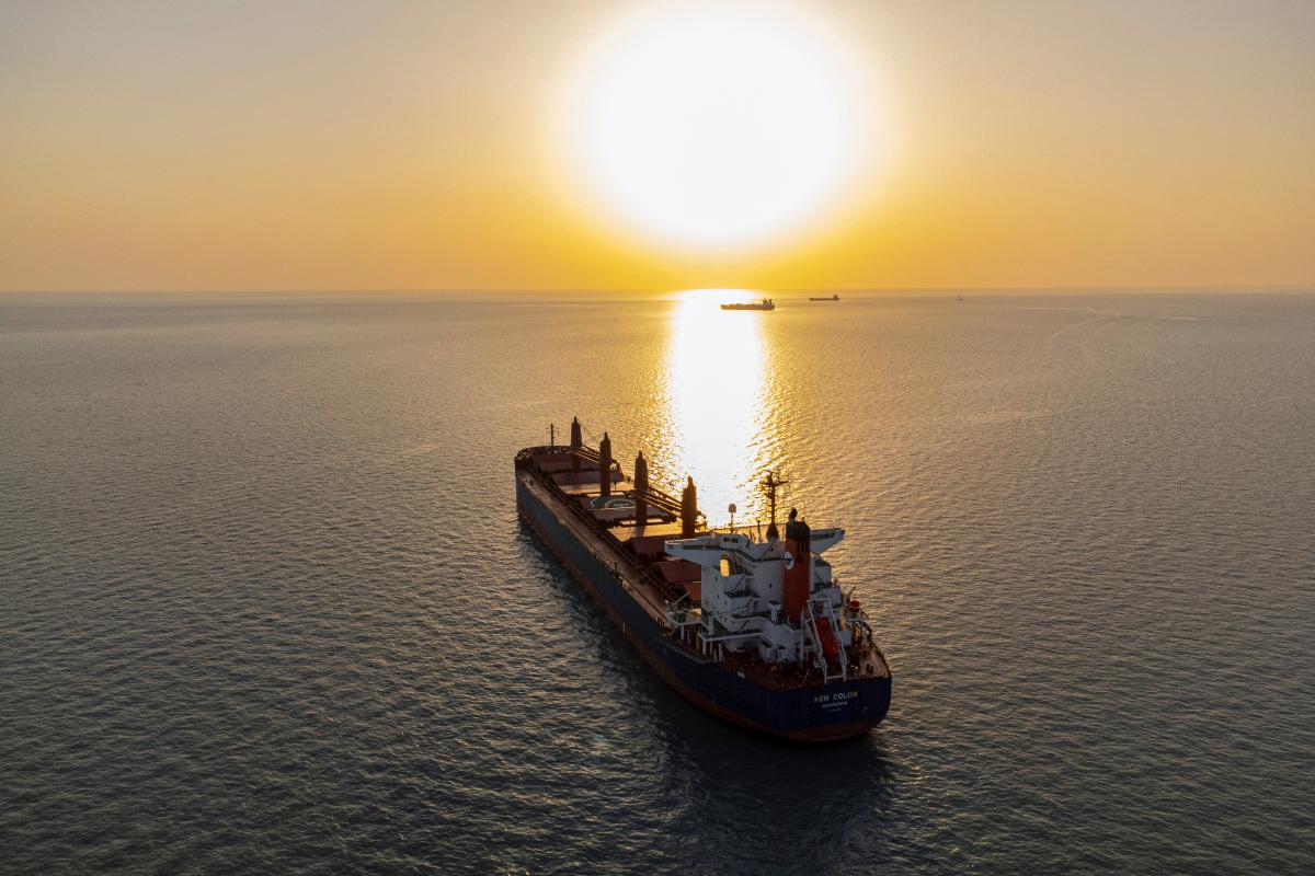 A cargo ship rests on the open ocean as the sun sets behind it, glinting off the sea surface