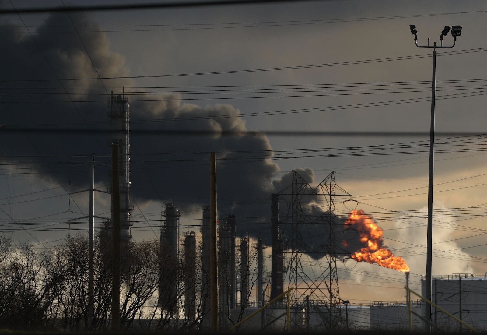 Smokestacks of a petrochemical refinery in the distance, with flames and smoke billowing out of one and blowing leftward.