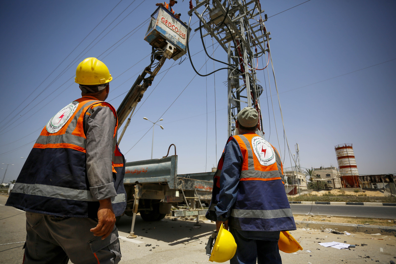 two people in construction hats and vest look at an electrical tower with construction equipment reaching up to it