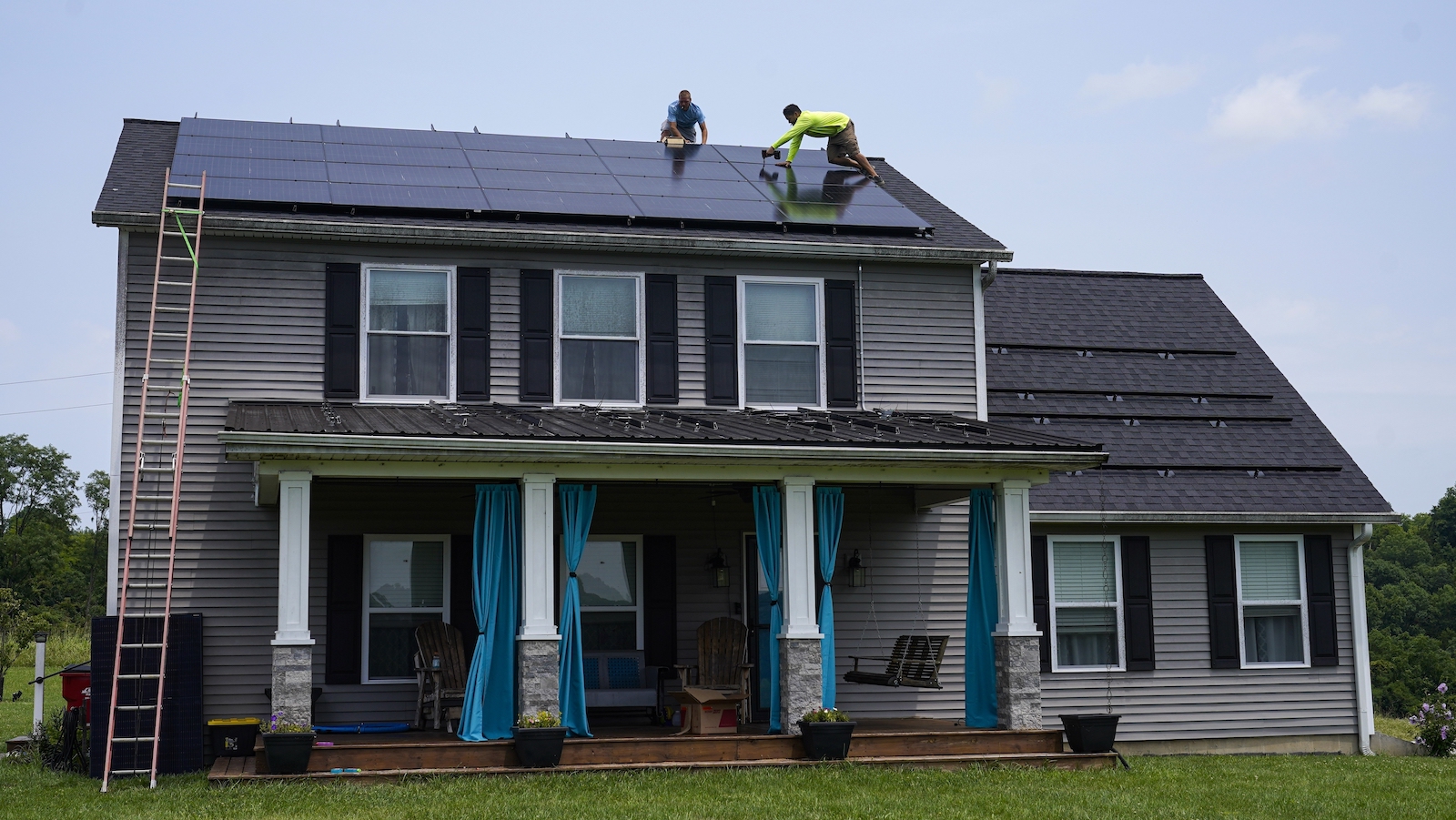 Two men install solar panels on the roof of a two-story house with a shingled roof and white columns on the gabled front porch.