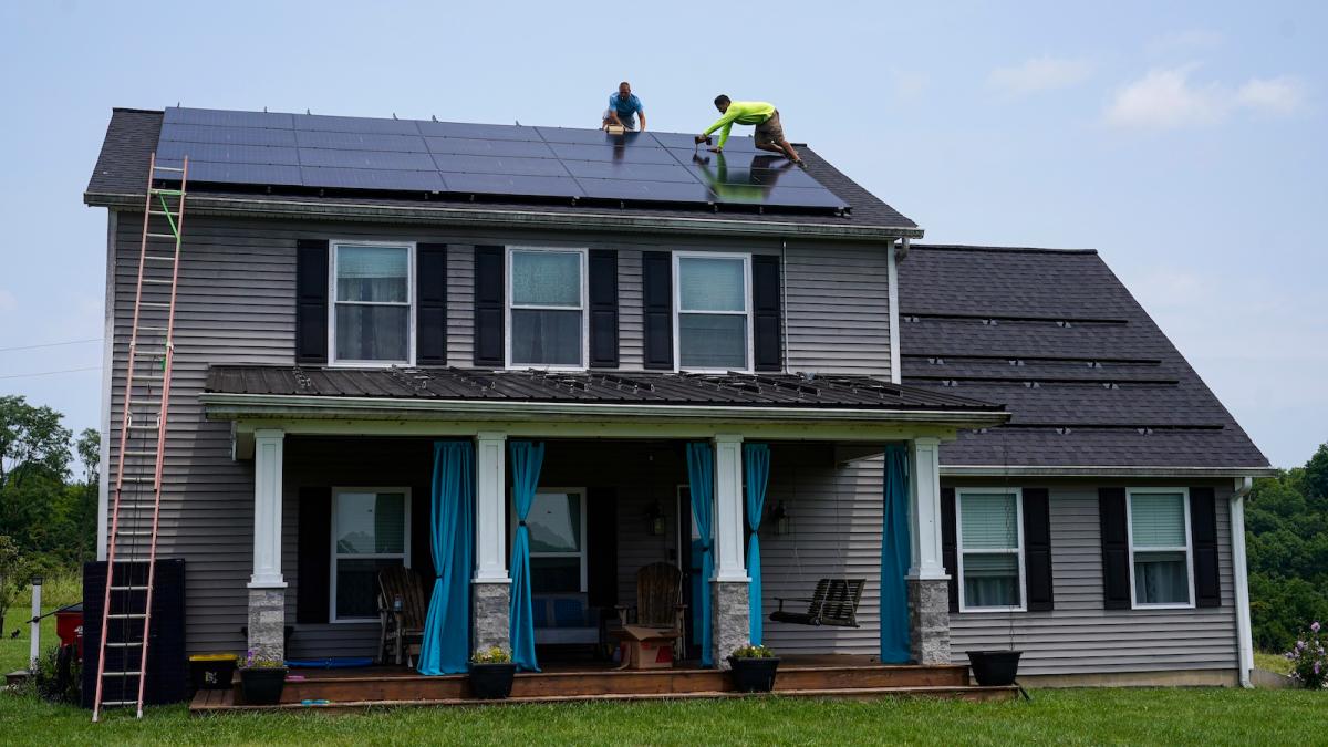 Two men install solar panels on the roof of a two-story house with a shingled roof and white columns on the gabled front porch.