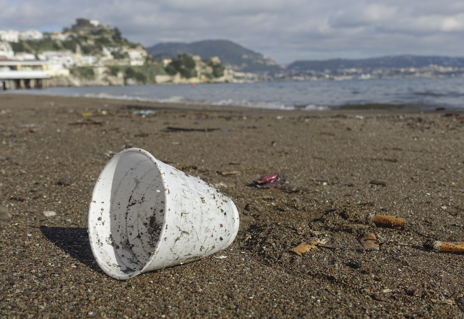 An empty plastic cup sits on its side on a beach, with water and buildings in the background.