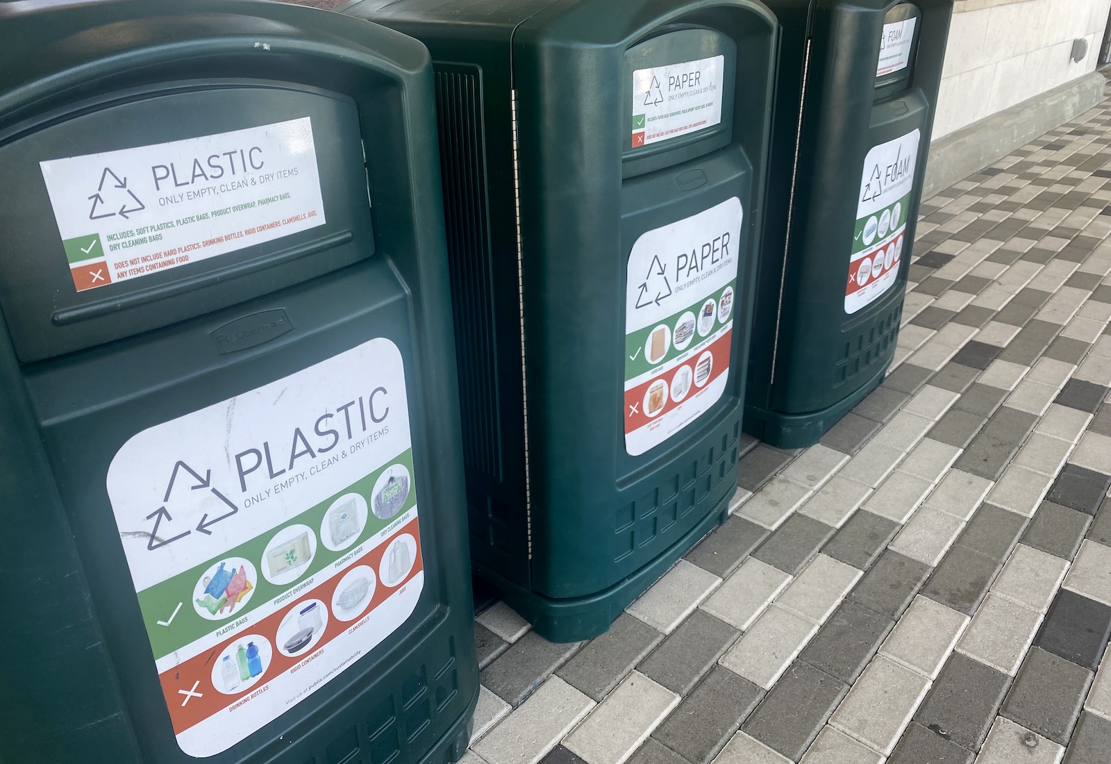 Three green recycling bins for plastic, paper, and foam in a row on a tiled floor. The bins have images on the front of them depicting recyclable items.