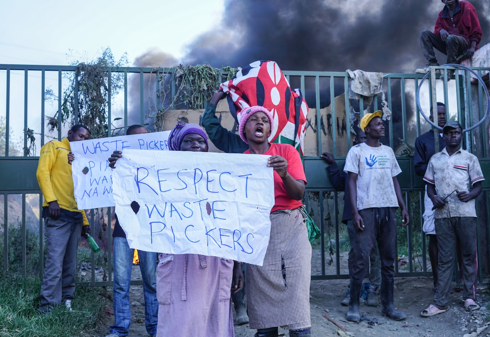 Two waste pickers in the foreground hold a sign reading, "Respect waste pickers." They stand in front of a fence, and smoke billows in the background.