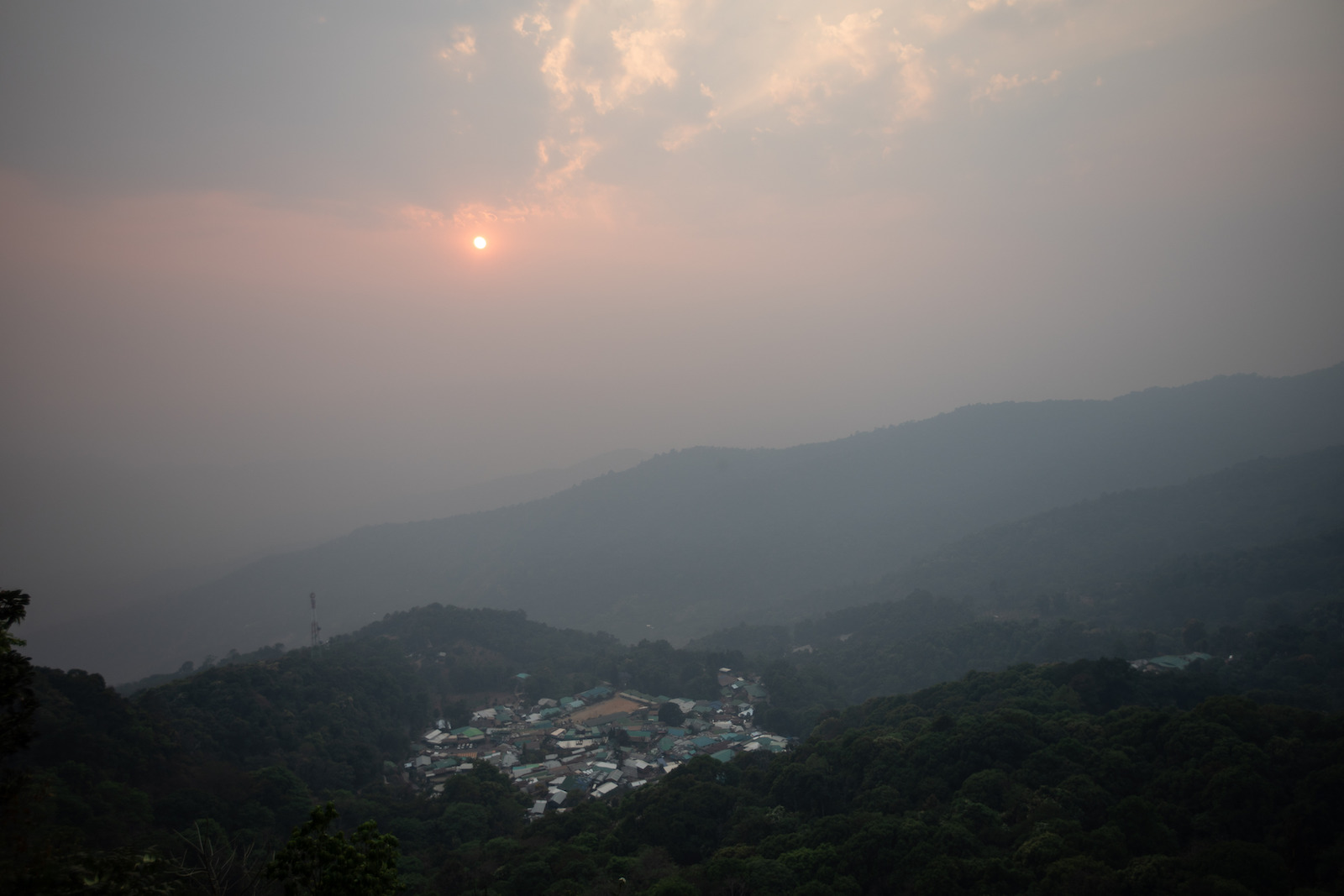 the sun is barely visible through a thick cloud of smoke over a mountain village