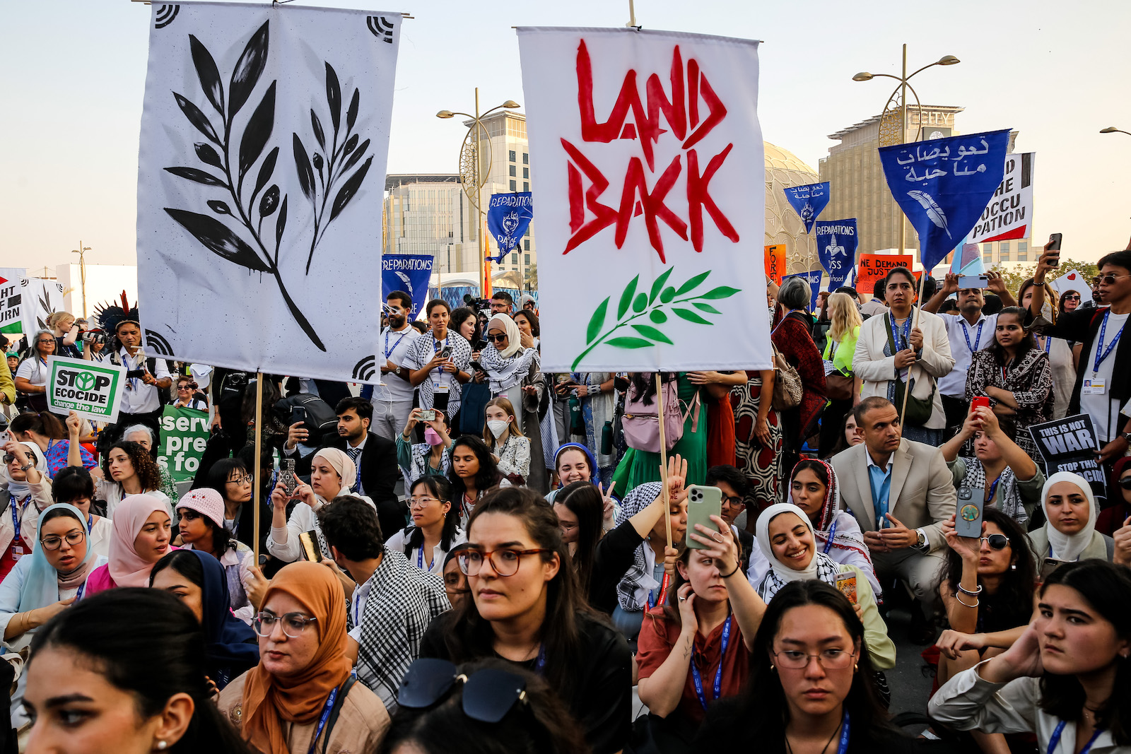 A large group of people hold signs with symbols and words including olive branches, arabic lettering, and 'land back' on them