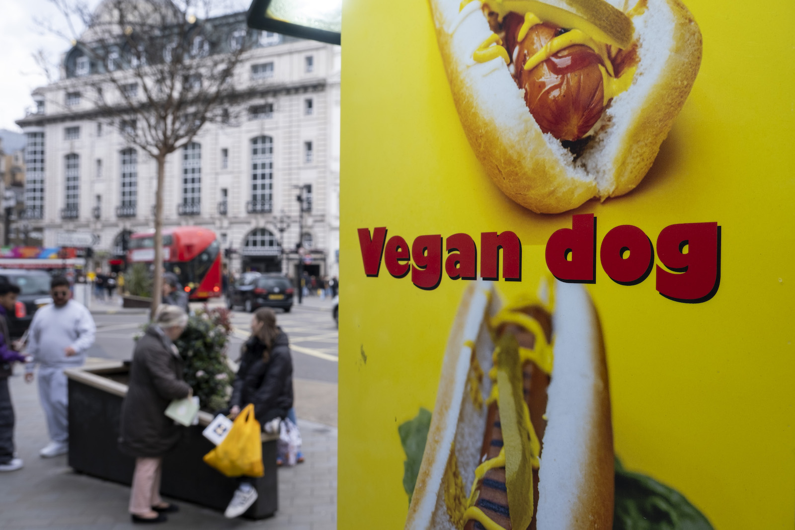 A yellow sign featuring the words 'Vegan dog' and photographs of hot dogs in the foreground of a gray urban scene