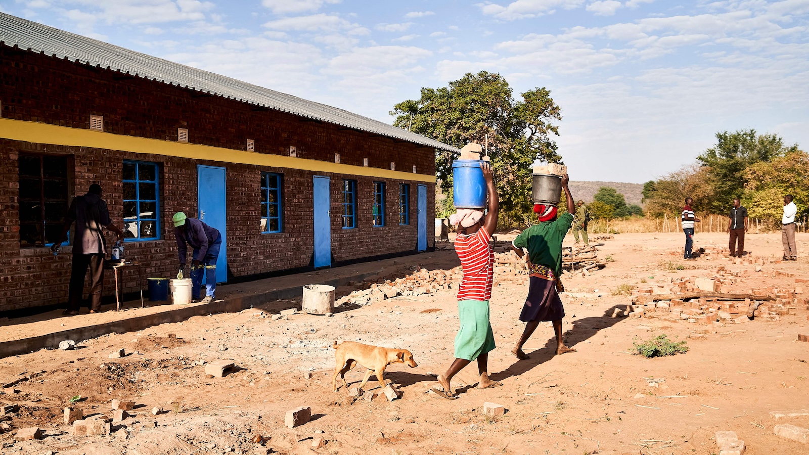 Two women carry buckets of bricks on their heads as they walk across the dirt. A brick building is to their left and trees are in the background.