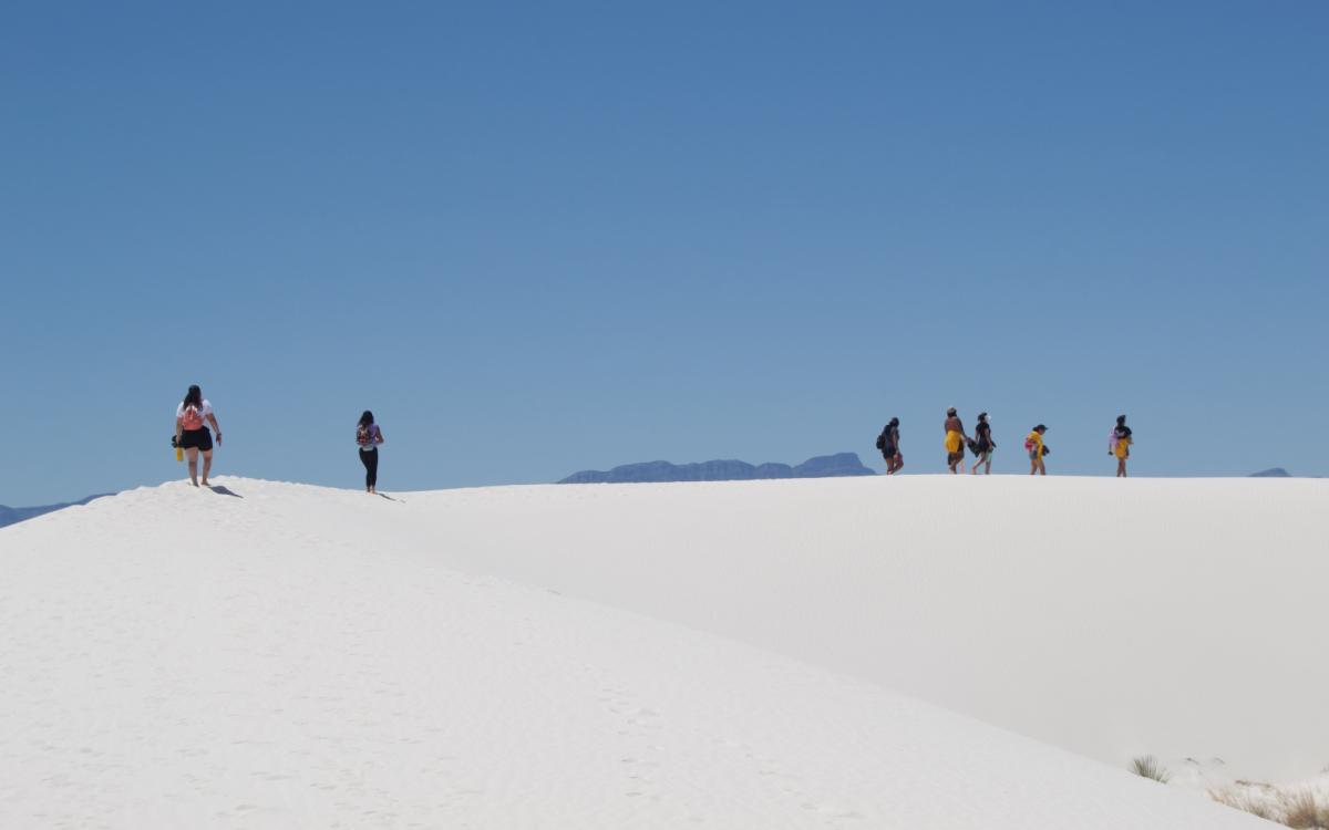 A group of about seven young people are walking along a bright white sandy dune with blue sky above