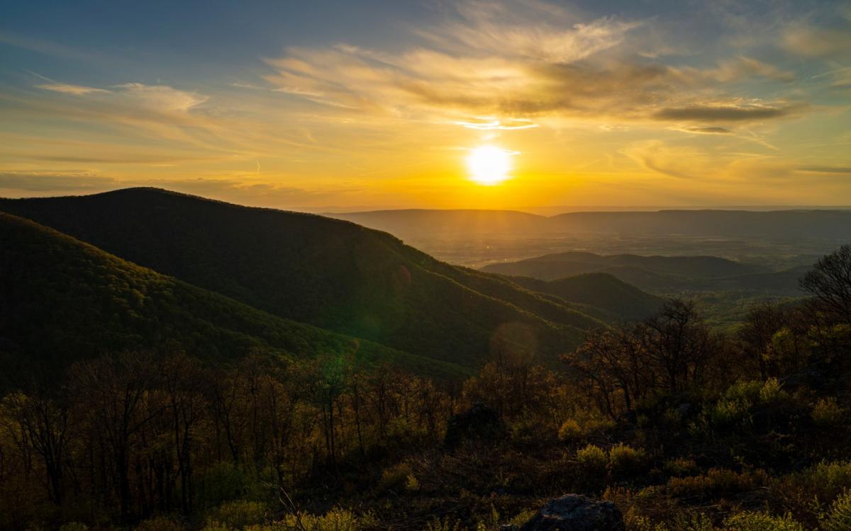 A golden sunset peaks over the horizon of tree-covered mountains