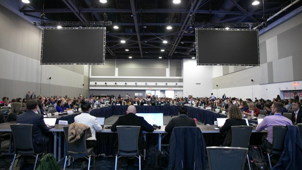 Wide shot of a room full of negotiators sitting at tables in a rectangular formation. Two large, blank screens hang from the ceiling.