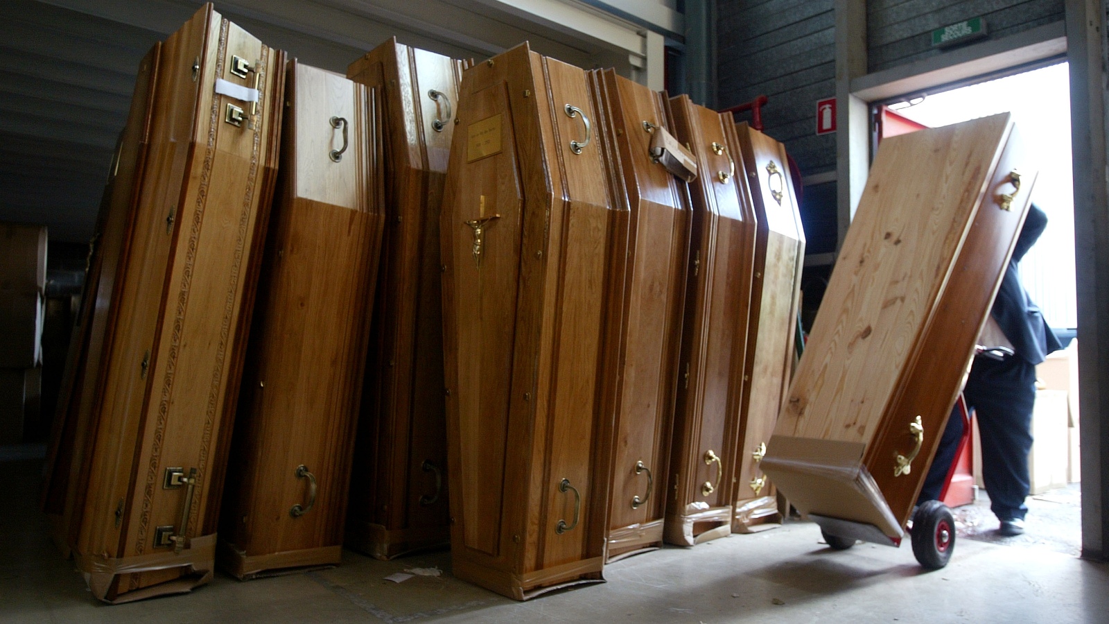 A man wearing a suit wheels a large brown wooden coffin into a room where similar-looking light-wooden coffins are stacked vertically.
