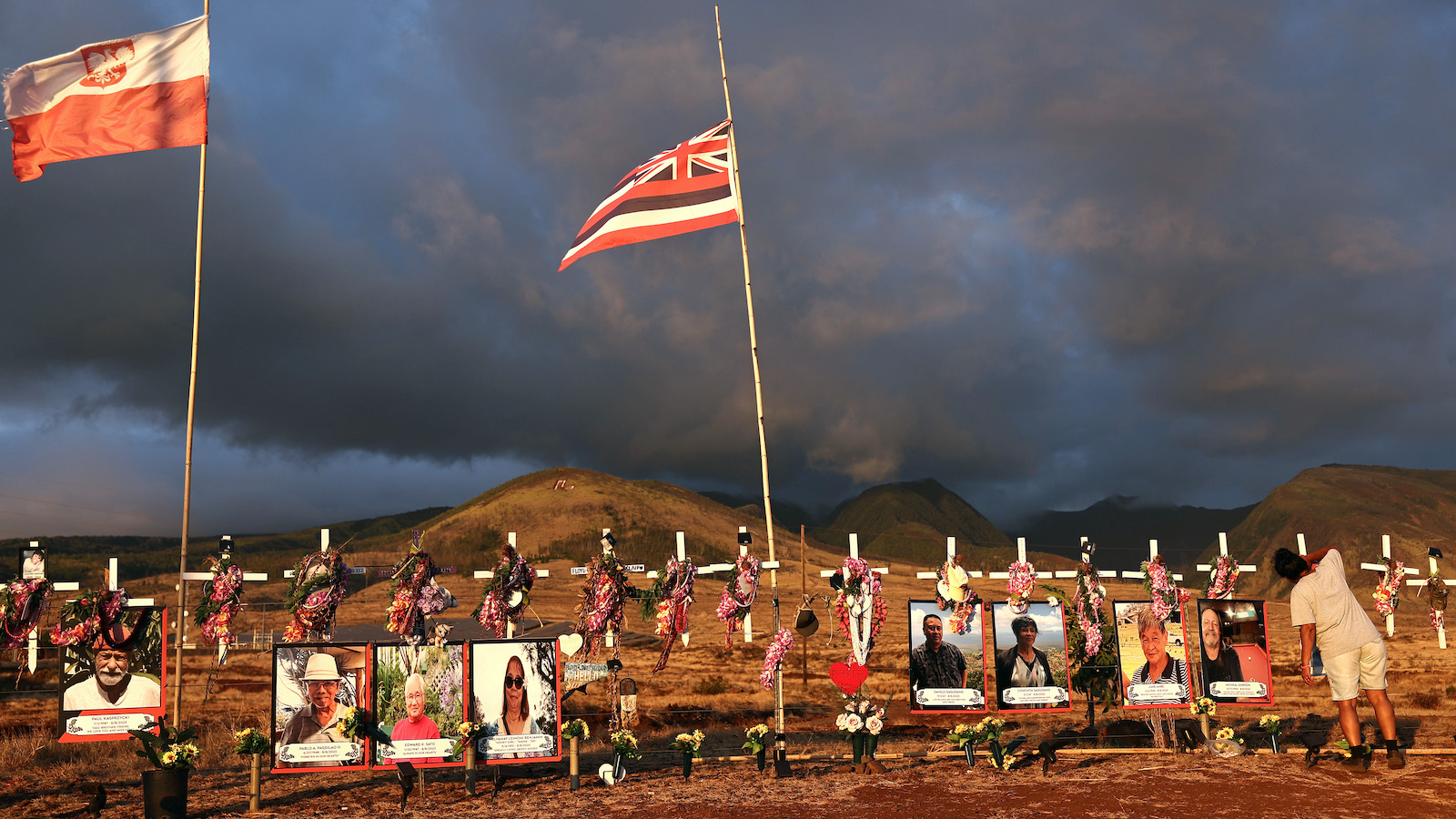 Dark sky and sand dunes in background of flags, photos and flowers in a memorial for victims of first anniversary of Lahaina fire on Maui.