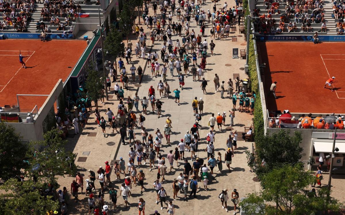 An aerial shot of a crowd walking between two red clay tennis courts on a hot day