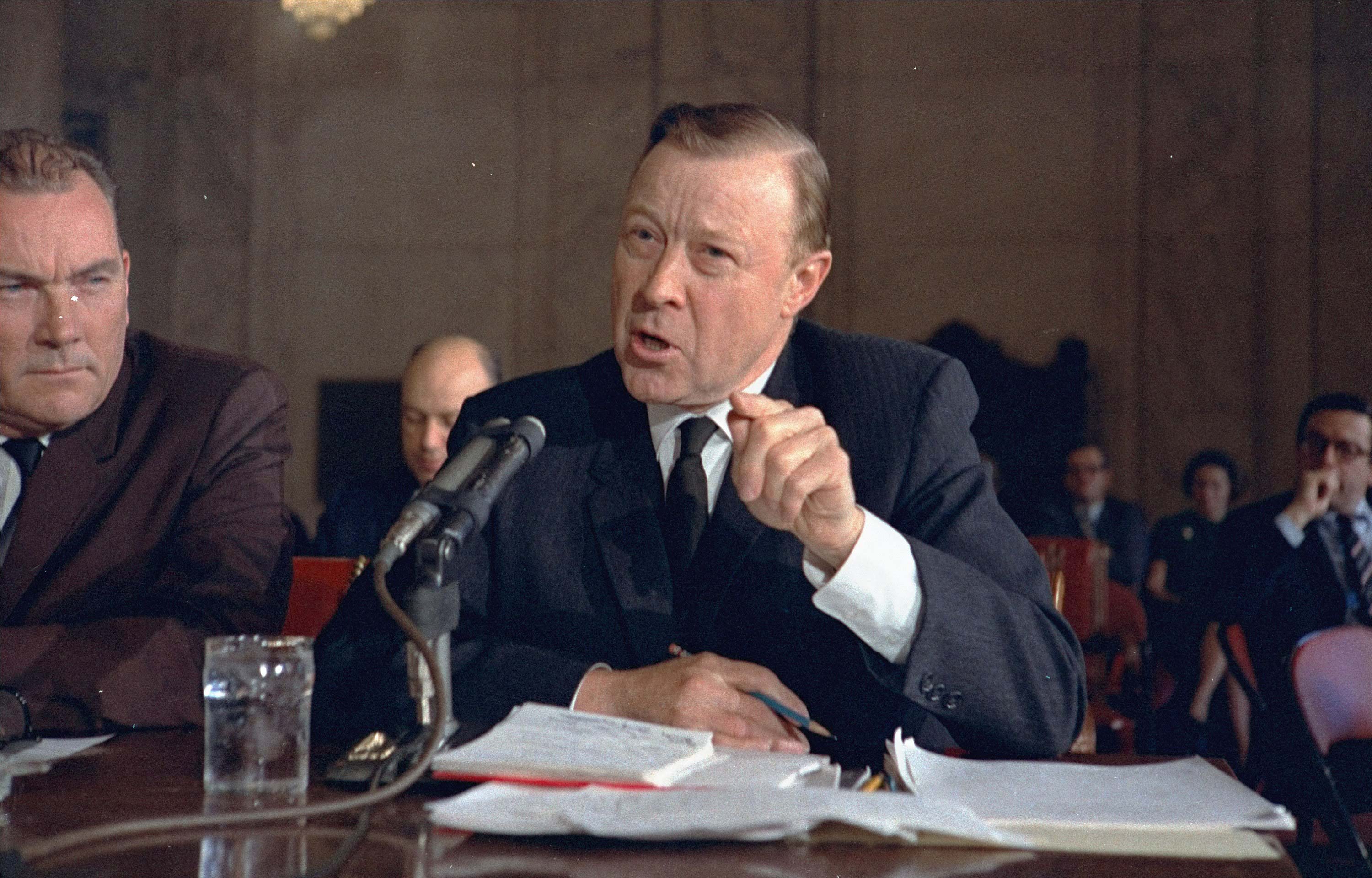 A man in a suit points forward while speaking in front of a microphone at a formal bench