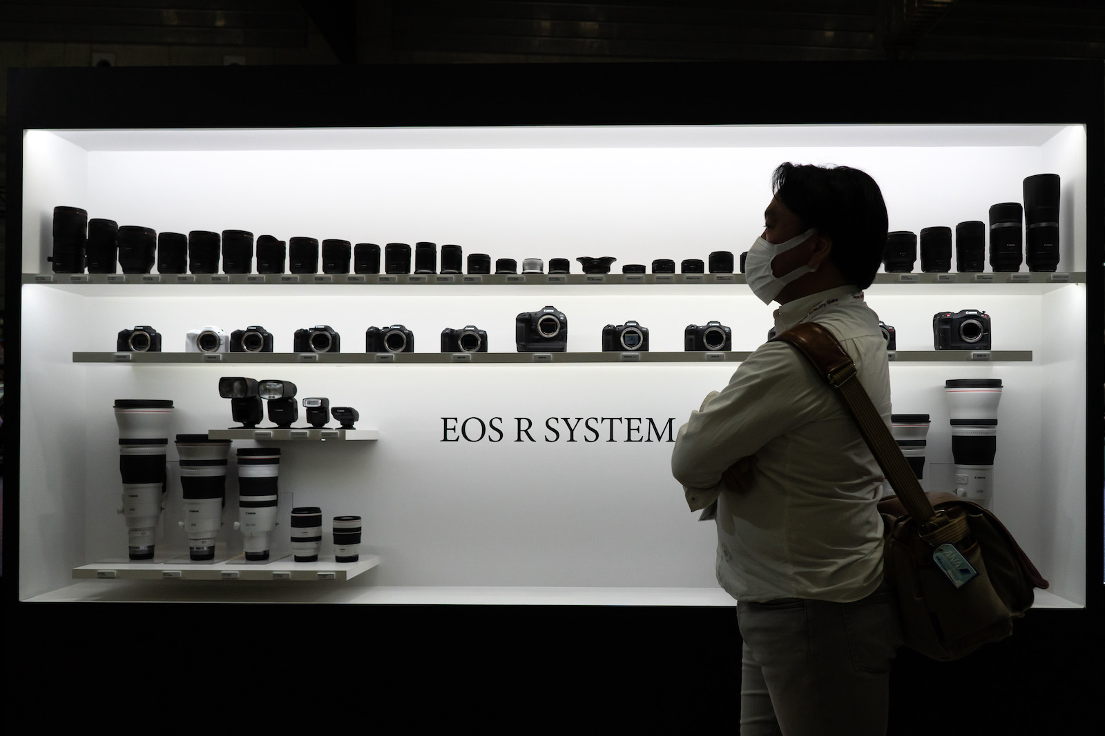 A person wearing a mask stands in profile in front of a sleek display of small cameras and equipment, which appear black against a white background bearing the text 'Eos R System'