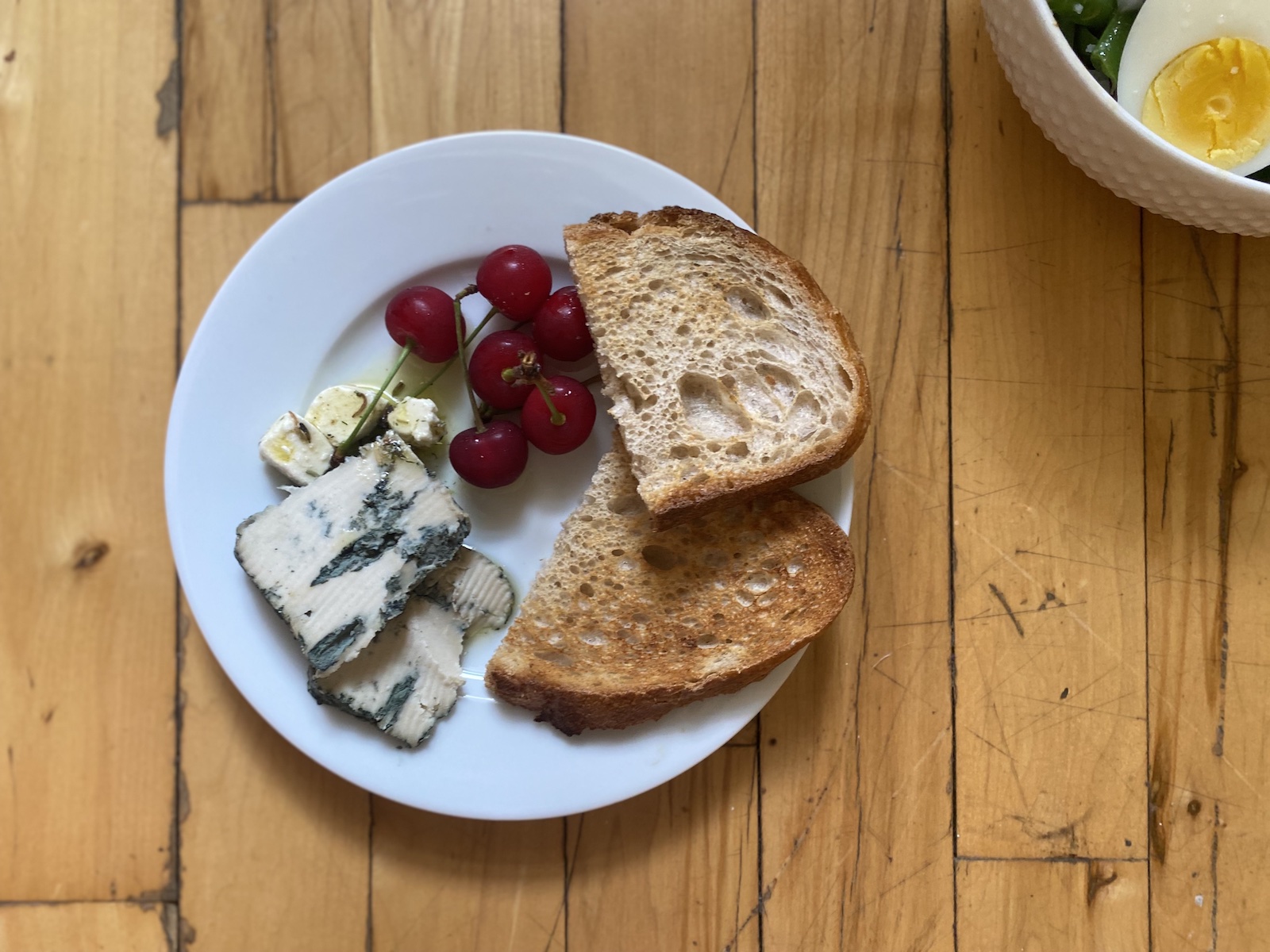 Overhead shot of a white plate with toasted bread, dark red cherries, two slices of blue cheese, and a few cubes of marinated feta cheese