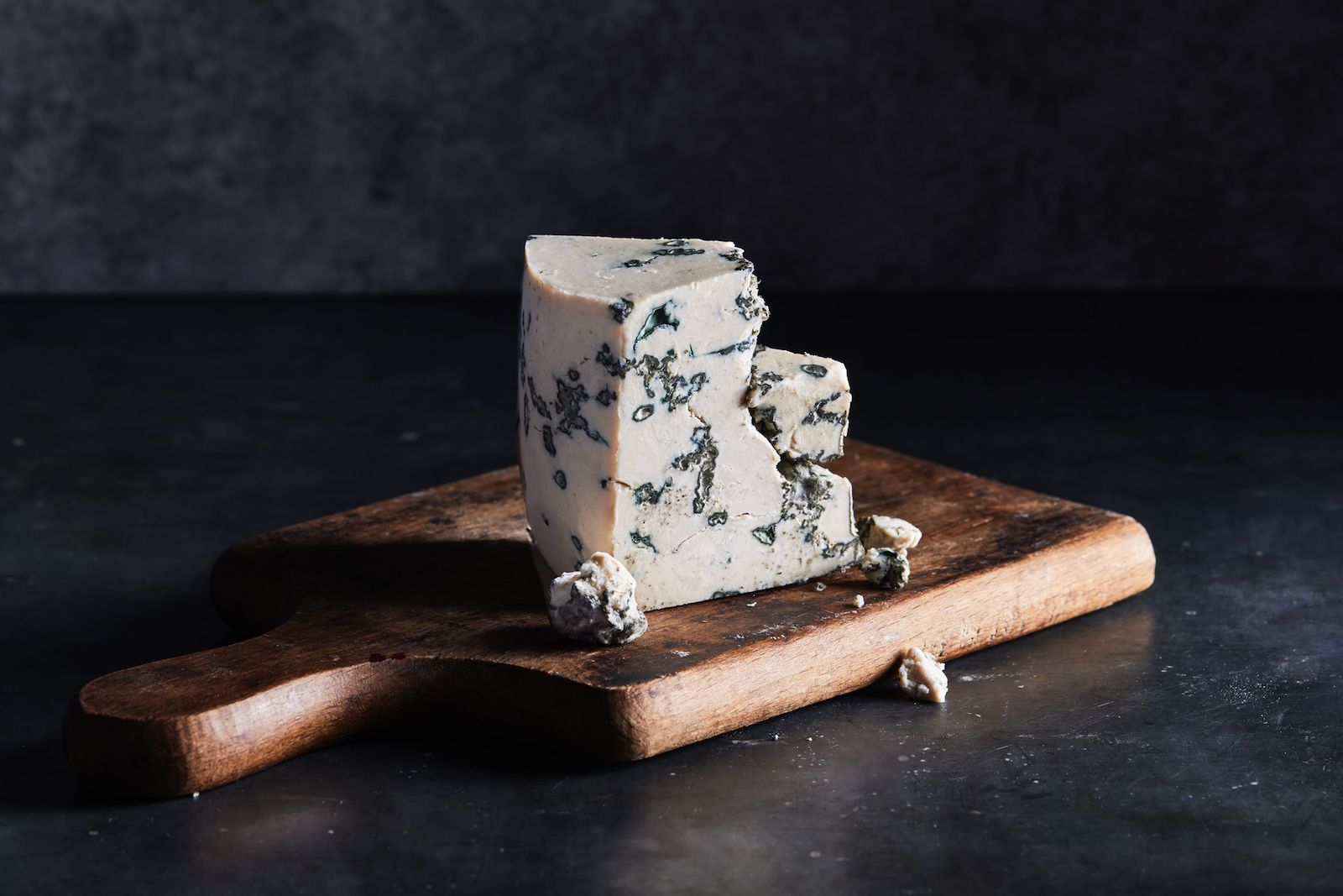 A wedge of blue cheese, with a few crumbles scattered around it, on a brown wooden cutting board against a dark background