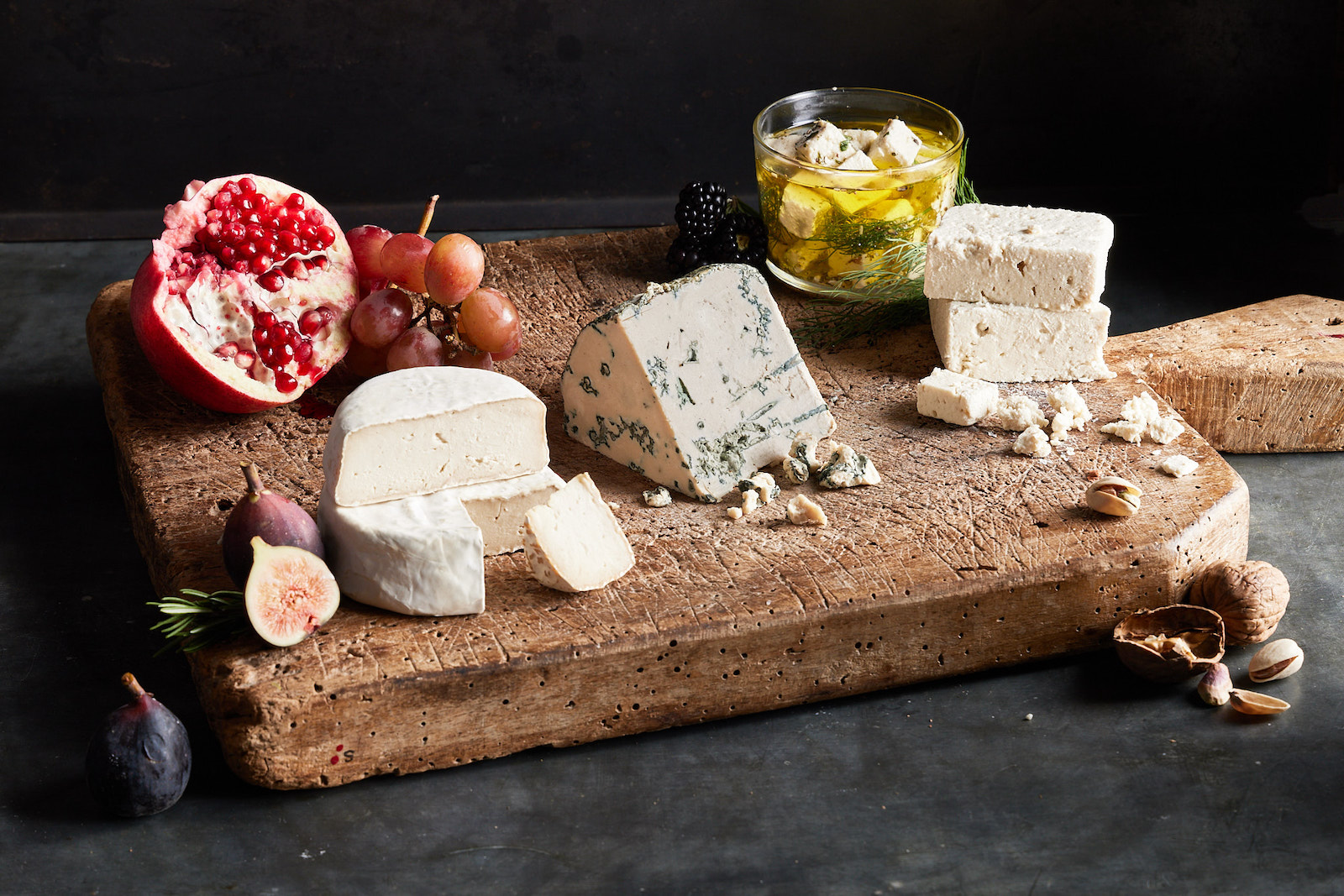 Hunks of brie, blue cheese, and feta sit on a well worn brown cutting board along with fresh figs, grapes, blackberries, and half a pomegranate