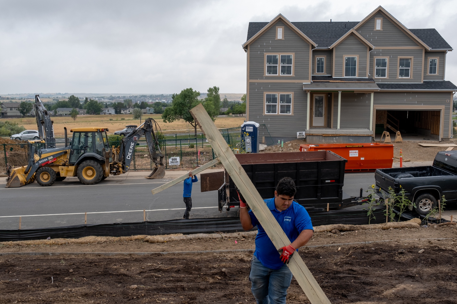 Two construction workers, one in the foreground and one in the background, haul wood planks in a neighborhood under reconstruction