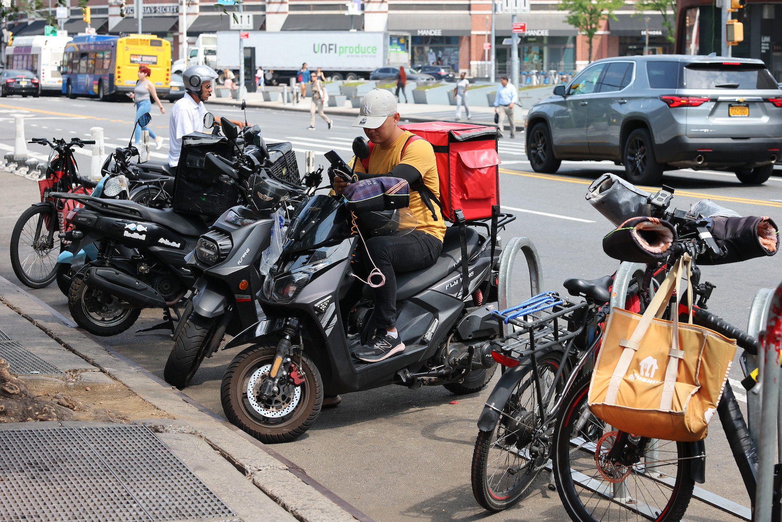 'n Persoon in 'n oranje hemp sit op 'n swart brommer met 'n rooi koeler op die rug, omring deur ander fietse en brommers, met 'n stadsstraat in die agtergrond