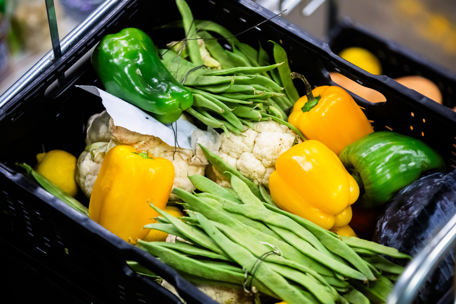 A black crate that contains bundles of long bean pods, yellow and green bell peppers, and white cauliflower