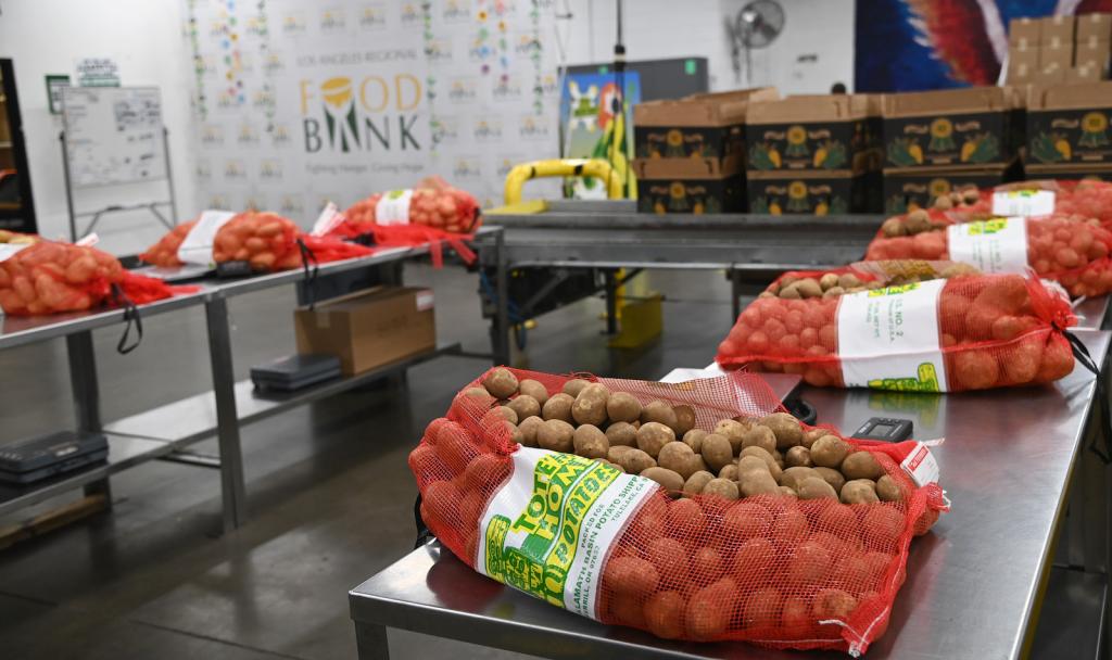 A large industrial room with a 'Food Bank' sign against the wall contains long metal tables with six very large red mesh bags of brown potatoes on them