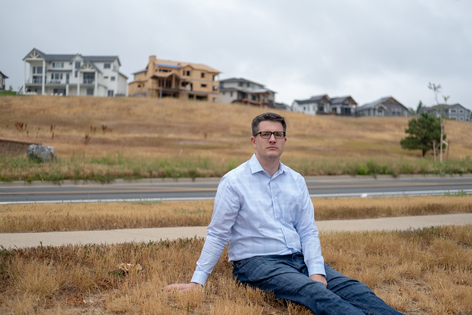 A man in glasses, a button-down shirt, and jeans sits in a grassy field in front of an under-construction housing development