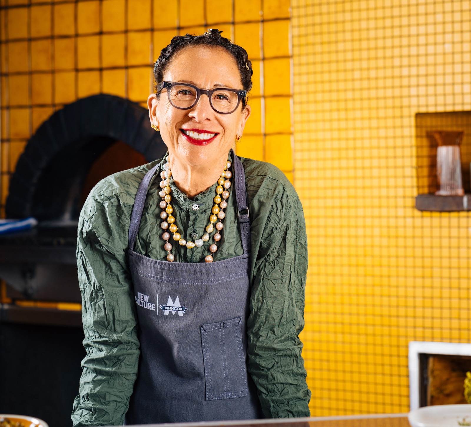 A smiling woman wearing gray-rimmed glasses, red lipstick, cream-colored beads, and a blue apron stands against a yellow background that includes a pizza oven