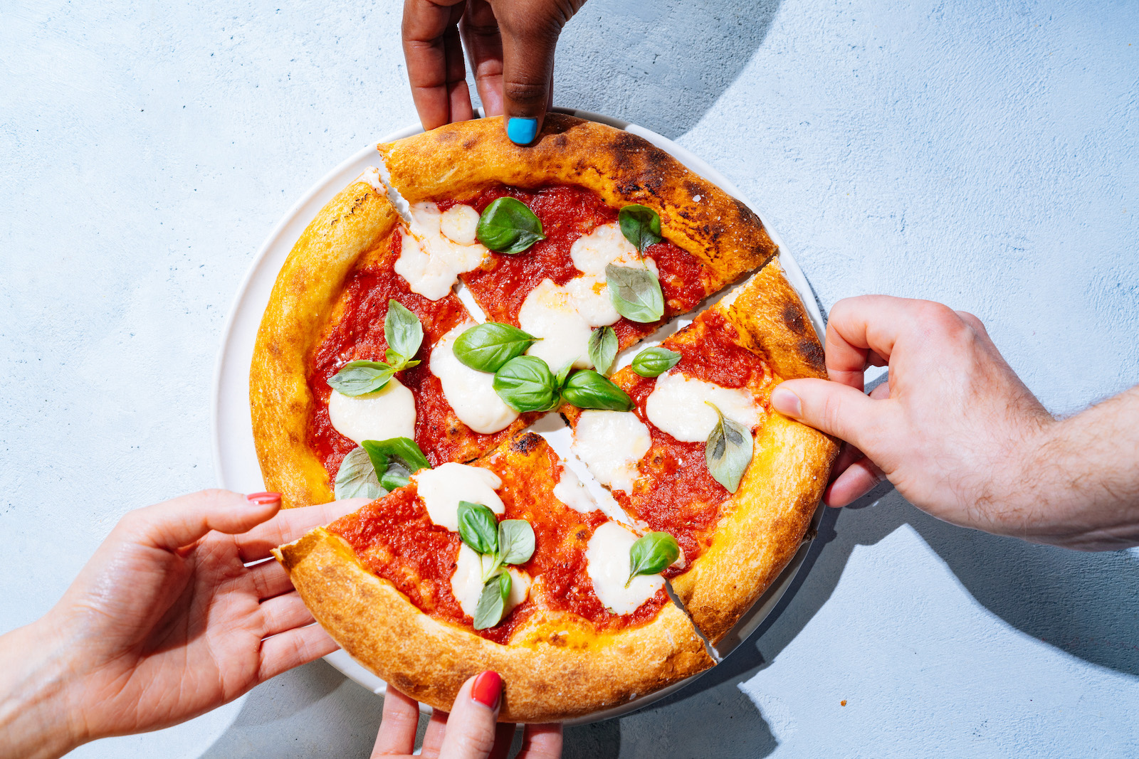 An overhead shot of a margherita pizza cut into quarters, with white cheese and green basil on top of a crust coated in red sauce, with four hands each slightly lifting one of the quarters