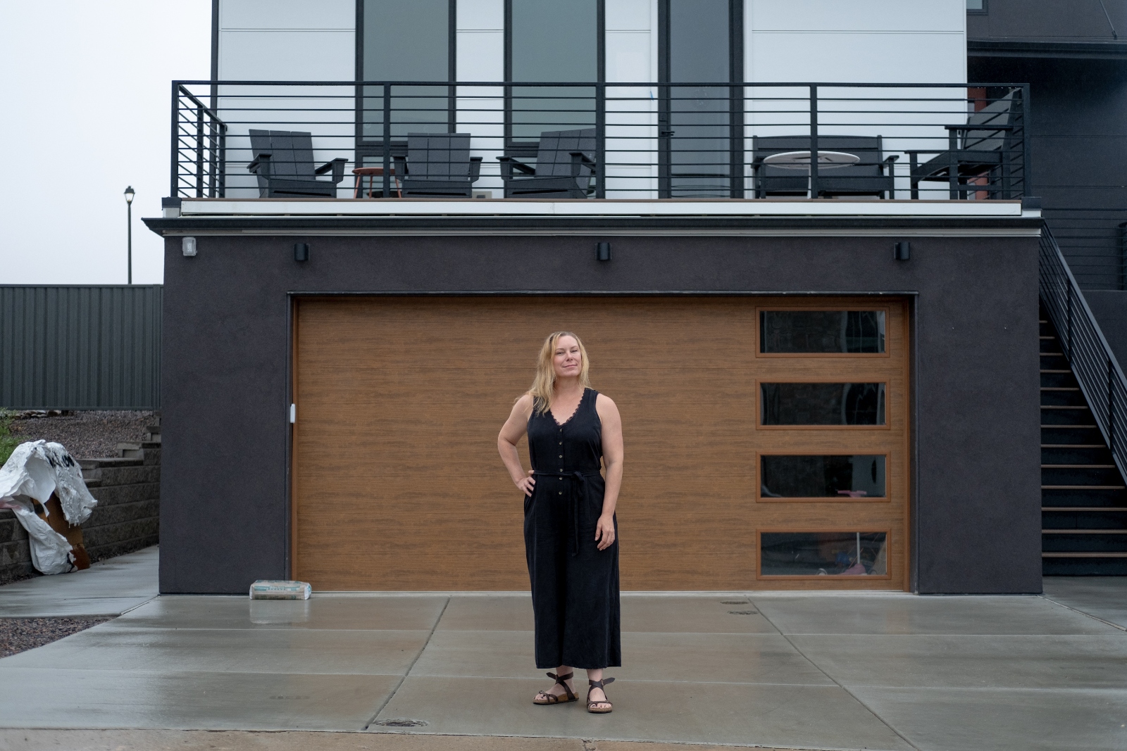 A woman wearing a long black dresses poses with her hand on her hip in front of a newly constructed house