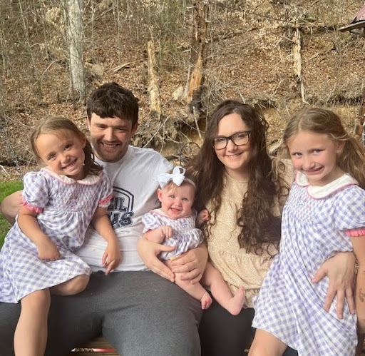 A man and woman sit in front of trees in the winter with two little girls and a baby.