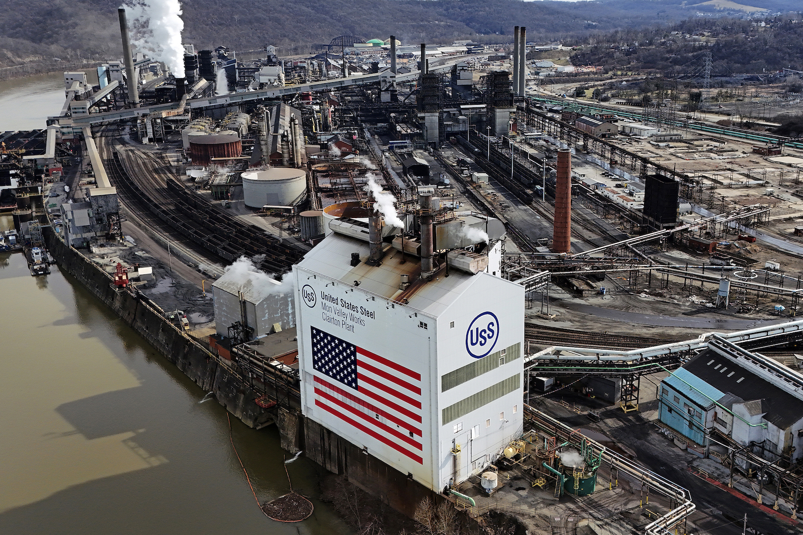 A large industrial facility near a river. In the foreground there is a white building with an American flag and the words United States Steel, Mon Valley Works, Clairton Plant