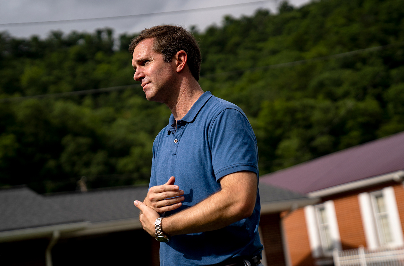 A man in profile looks before him and folds his arms. The man is Kentucky Governor Andy Beshear and he is talking to the press.