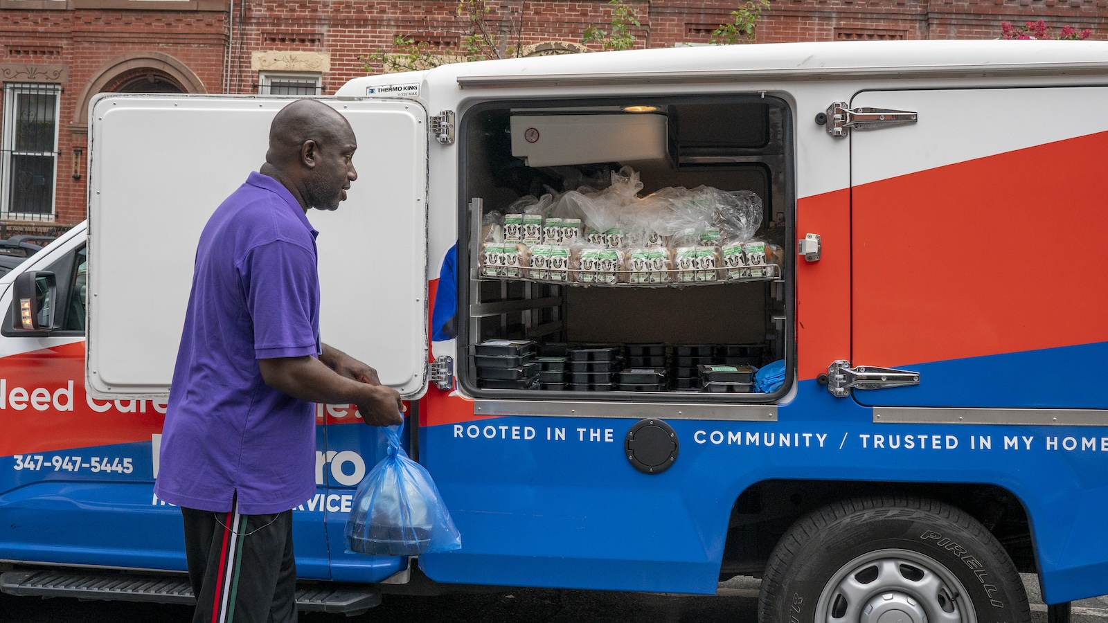 A man in a purple shirt stands in front of a van with its doors open, revealing a shelf of bread