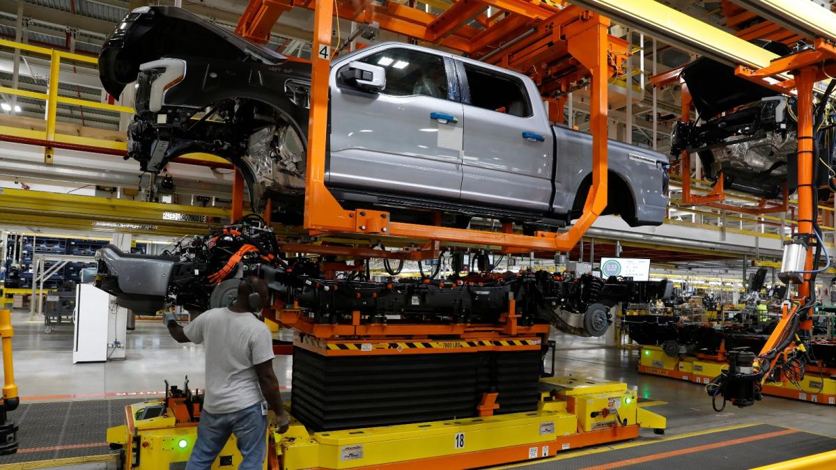 A man in a tee shirt and jeans raises a pickup truck on a piece of orange machinery in a factory