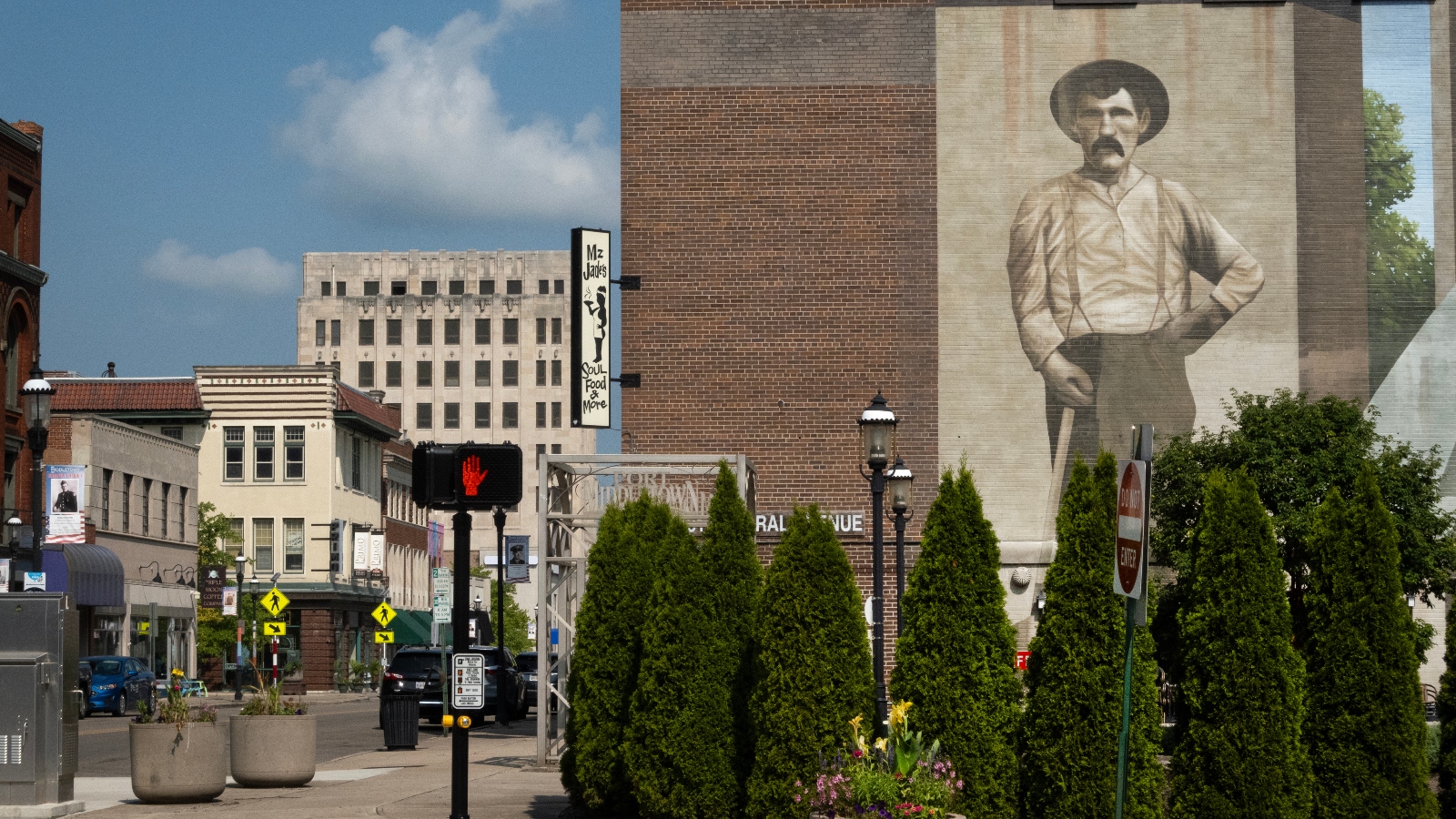 A mural of an old man wearing a hat stands on a brick building on a small town street.