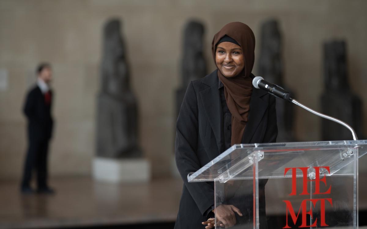 A young woman with a headscarf smiles, standing in front of a podium with the label "THE WITH" with a microphone